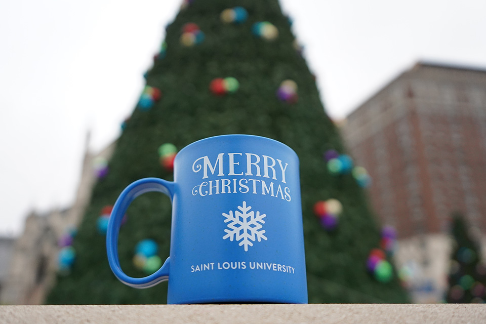 COTQ mug A mug says "Merry Christmas Saint Louis University" with an image of a snowflake on it as it rests in front of a large Christmas tree outside.