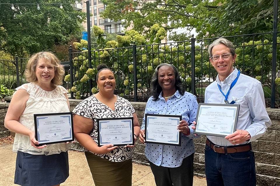 Four people hold awards plaques while standing under a tree.
