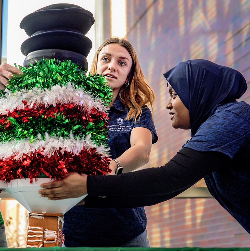 Two students hold the bottom of a stack of weights on top of a gingerbread house from the side to balance it.