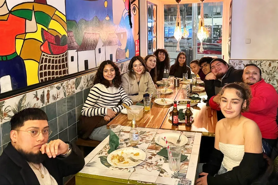 Several people are gathered around a table with plates of food on them as they smile for the camera. There is artwork on the right wall and a painted mural on the left. 