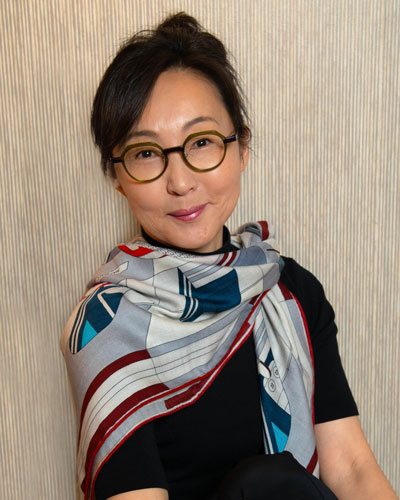 Janet Park Performance artist Janet Park smiles wryly at the viewer. She wears a black shirt with a scarf bearing geometric patterns in grays, blues, and reds.