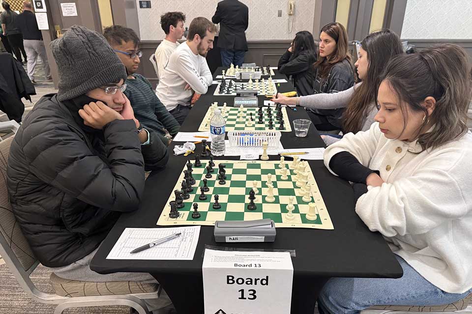Two chess players focus intensely on the game at Board 13. A man in a beanie and a woman in a white sweater analyze the chessboard. The atmosphere is serious.