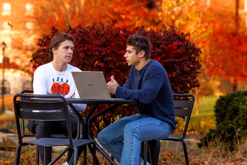 Two male students site at a table outside with a laptop
